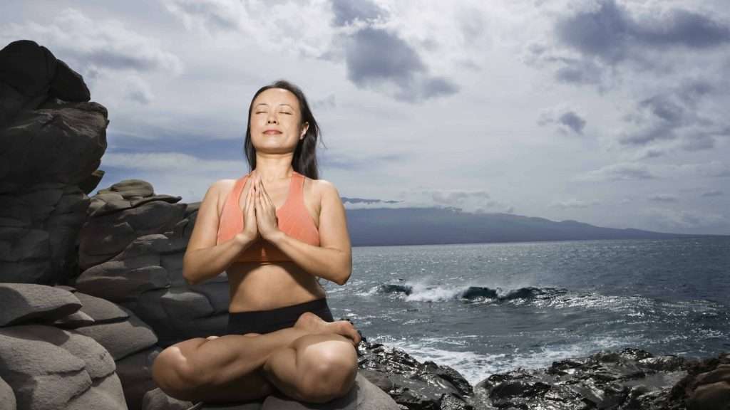 Asian woman sitting on rock by ocean in lotus pose with eyes closed in Maui, Hawaii